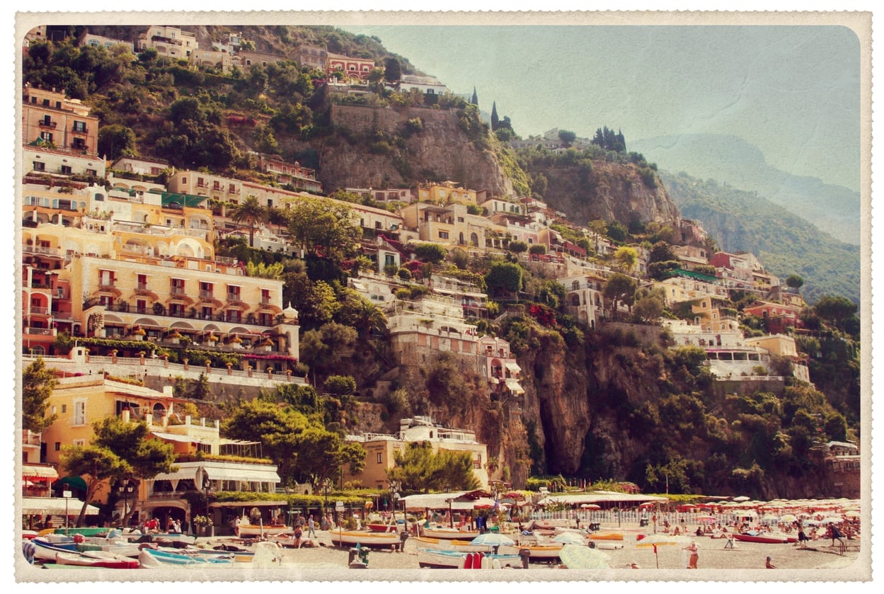Colorful hillside buildings overlook a busy beach with umbrellas and boats at the shore in Positano, Italy, on a sunny day. The image has a vintage, postcard-like appearance that evokes the charm of travel in the 60s and 70s.