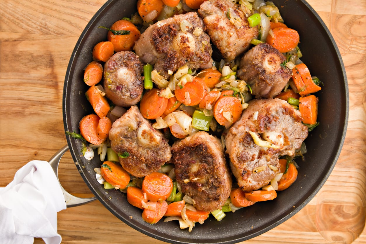 A pan of browned meat pieces with sliced carrots, celery, and onions sits on a wooden surface, ready to serve.