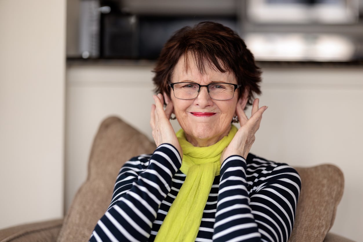 An older woman with glasses and a yellow scarf smiles while sitting on a couch, touching both sides of her face. She wears a black and white striped shirt and there is a blurred kitchen in the background.