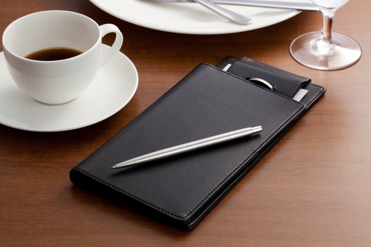 A black bill folder with a silver pen rests on a wooden table beside a cup of coffee, a white plate, cutlery, and a wine glass.