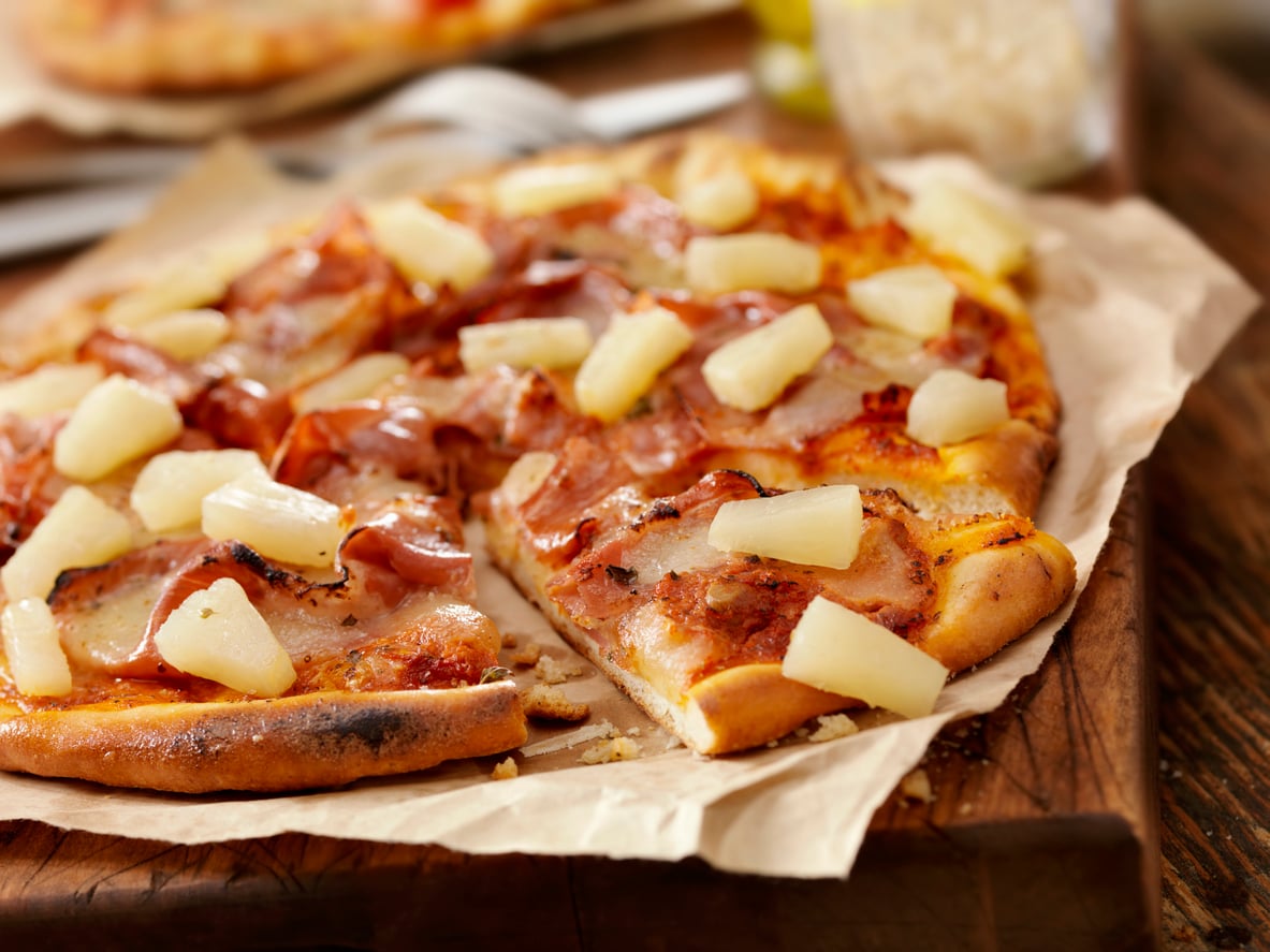 A Hawaiian pizza, one of the most beloved foods, is topped with ham and pineapple slices and sits on parchment paper on a wooden board, with one slice slightly pulled away from the rest.