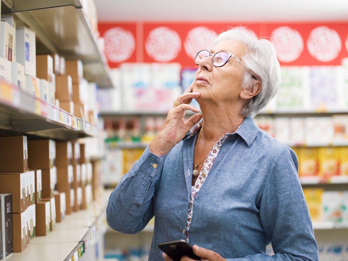 An older woman with glasses stands in a store aisle, holding a smartphone and looking thoughtfully at products on the shelves.