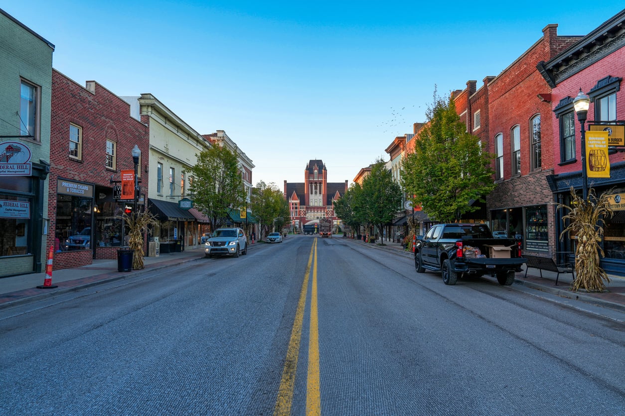 A quiet downtown street lined with shops and trees leads to a large historic building in the distance under a clear blue sky.