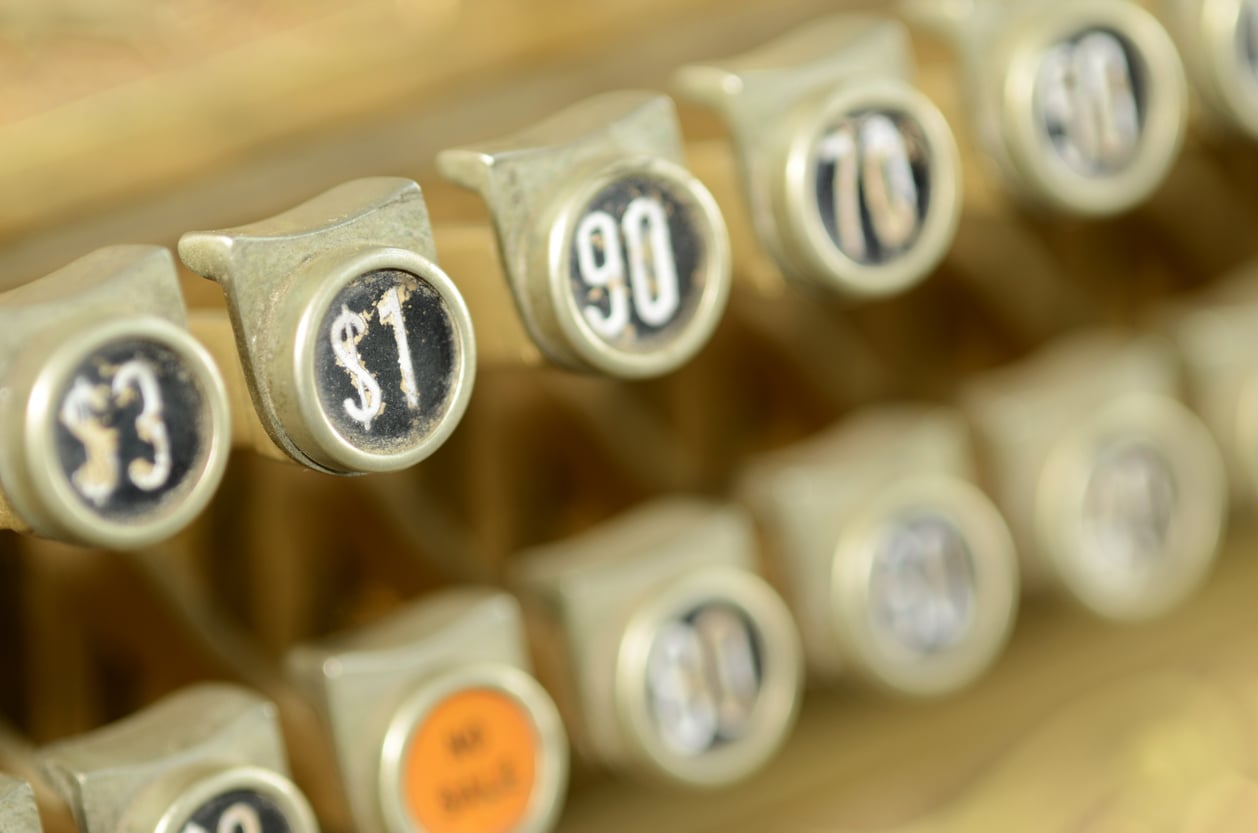 Close-up of vintage cash register buttons displaying various dollar amounts, including $1, $3, and $0.90, with worn metal and blurred background.