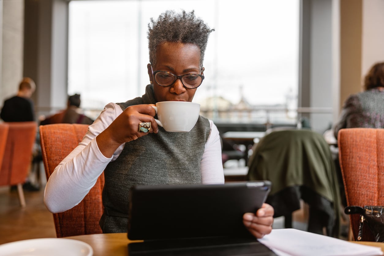 A person with short curly hair and glasses sits at a table in a cafe, drinking from a white cup while looking at a tablet. Other people are sitting in the background.