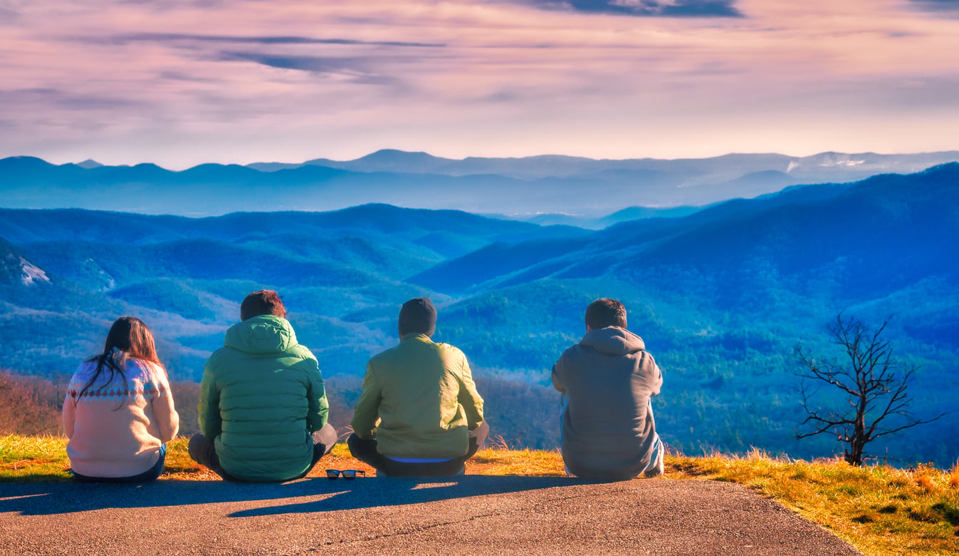 Four people sit on a grassy hill, facing a vast landscape of blue mountains under a colorful sky with soft clouds, enjoying the scenic view together—an ideal moment at one of the region’s best hiking destinations.