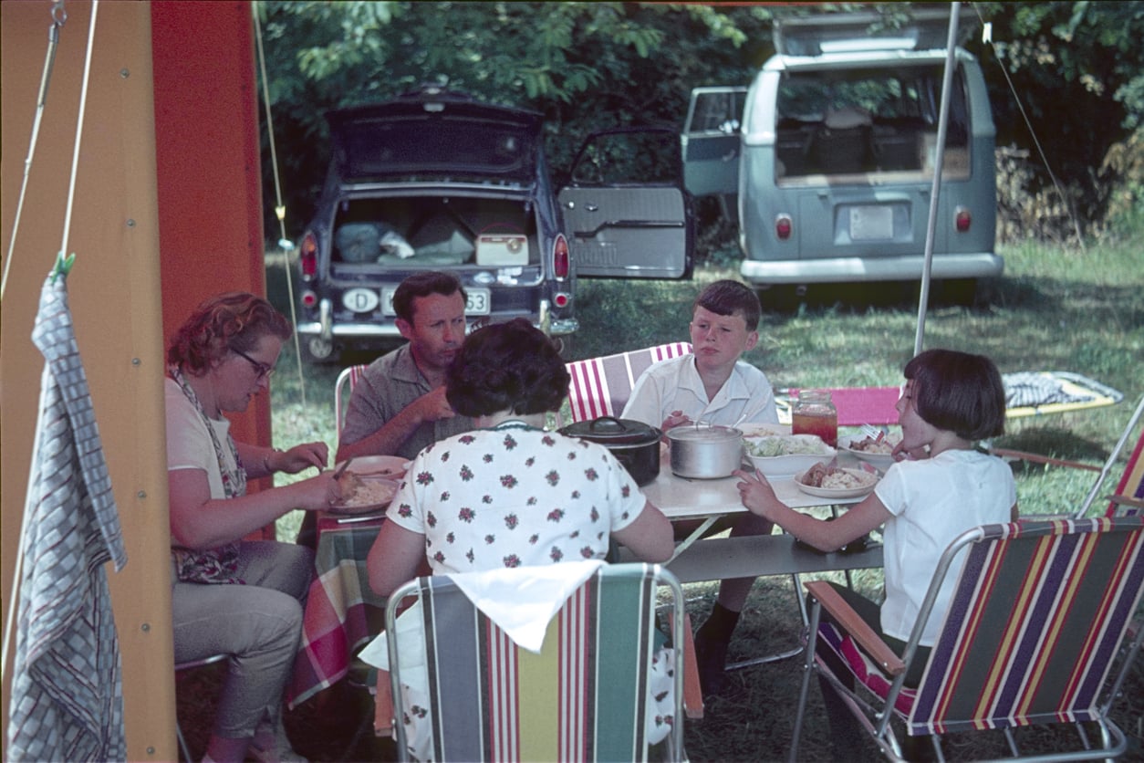 Five people sit around a table eating a meal at a campsite, with a car and camper van in the background—a classic scene reminiscent of travel in the 60s and 70s. The group gathers outdoors under a canopy, with striped chairs and trees behind them, evoking the relaxed charm of vintage road trips.