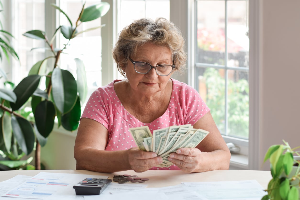 Older woman in glasses and a pink polka dot shirt demonstrates smart retirement money habits as she counts dollar bills at a table with papers, coins, and a calculator, near a window with green plants.