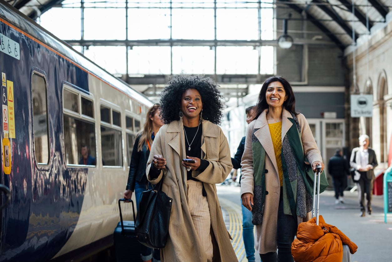 Two women smile as they walk on a train platform, each carrying luggage. Other people and a train are visible in the background inside a large, bright station.