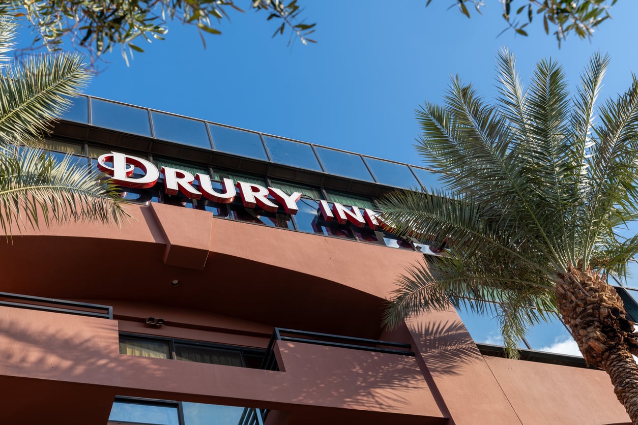 A low-angle view of a Drury Inn hotel building, one of the well-known hotel chains, with large signage, palm trees, and a clear blue sky overhead.