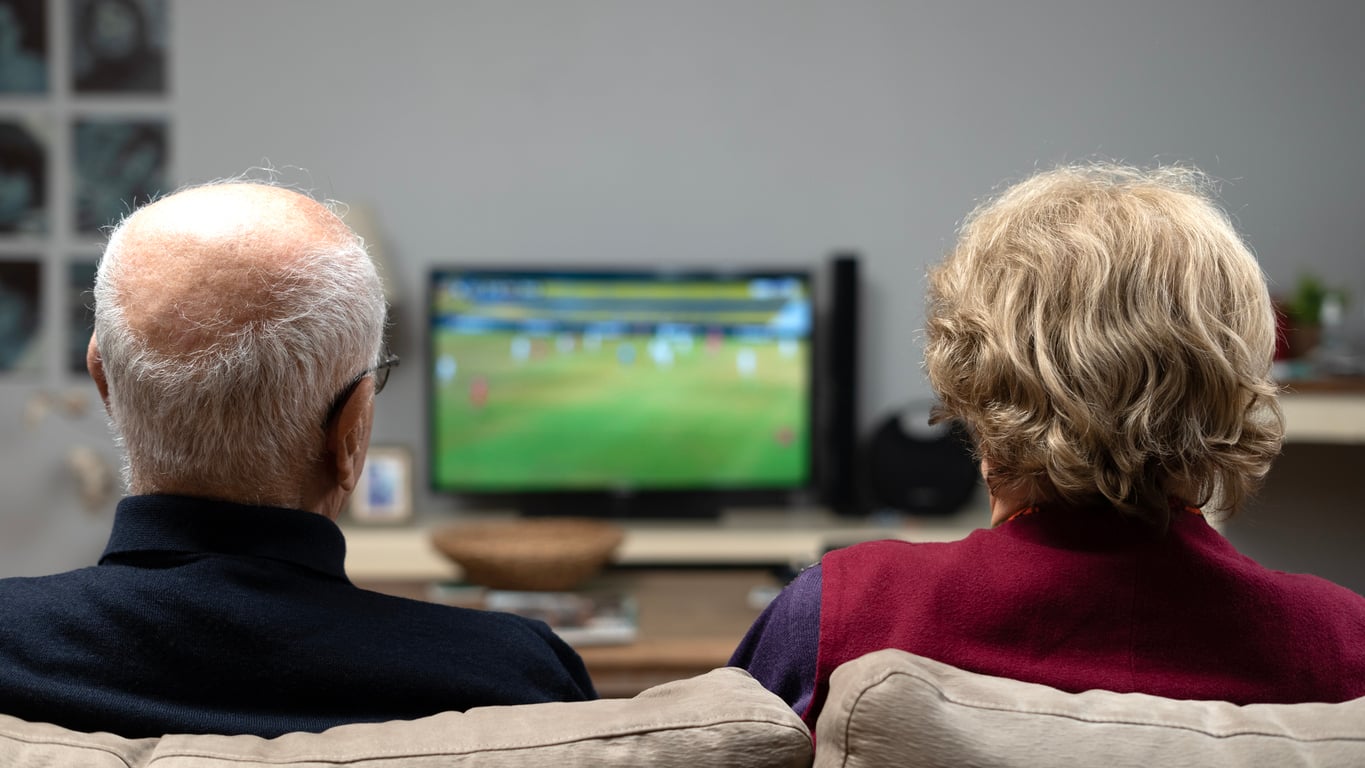 Two older adults sit on a couch, seen from behind, watching a soccer game on a television in a cozy living room setting.