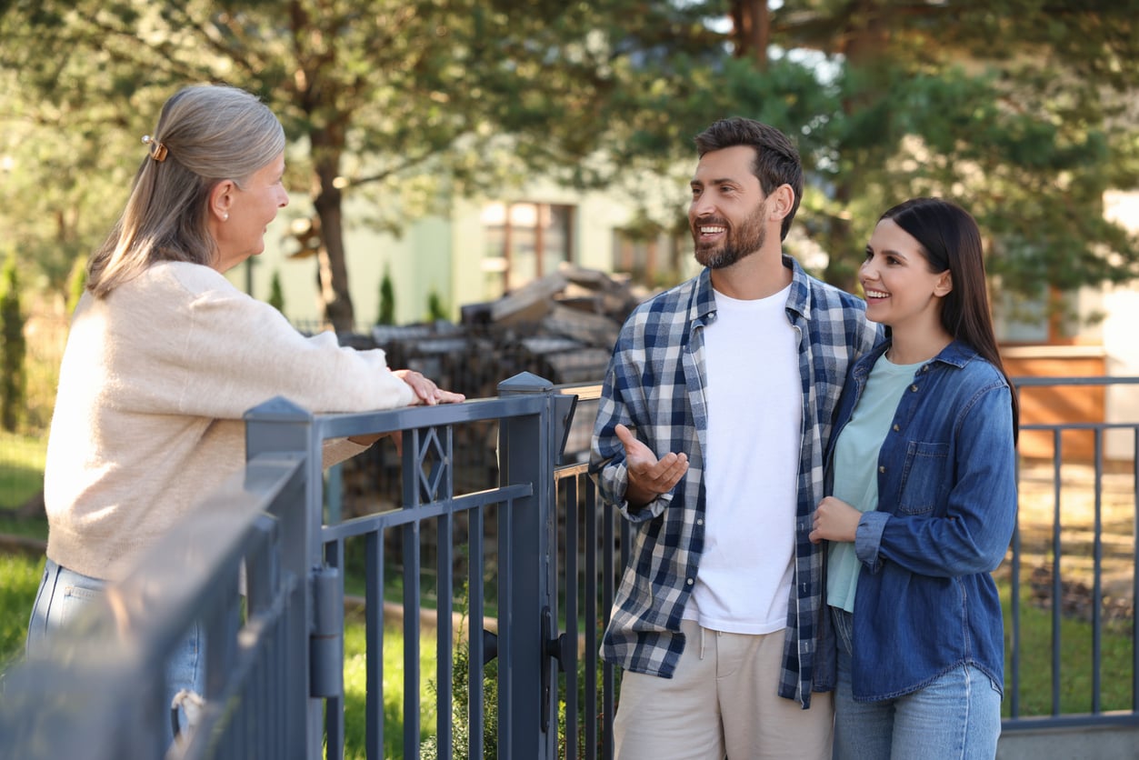 A smiling older woman stands by a fence, talking to a young couple outside on a sunny day. The couple appears happy and engaged in conversation, with trees and houses visible in the background.