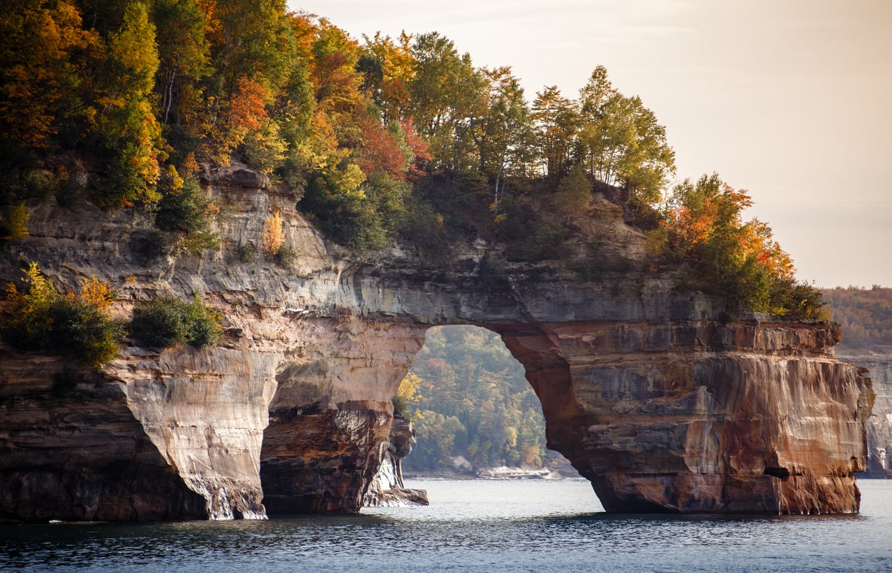 A natural rock arch rises over Lake Superior, topped with autumn trees showing red, orange, and green foliage under a cloudy sky—one of the region’s most breathtaking hiking destinations.