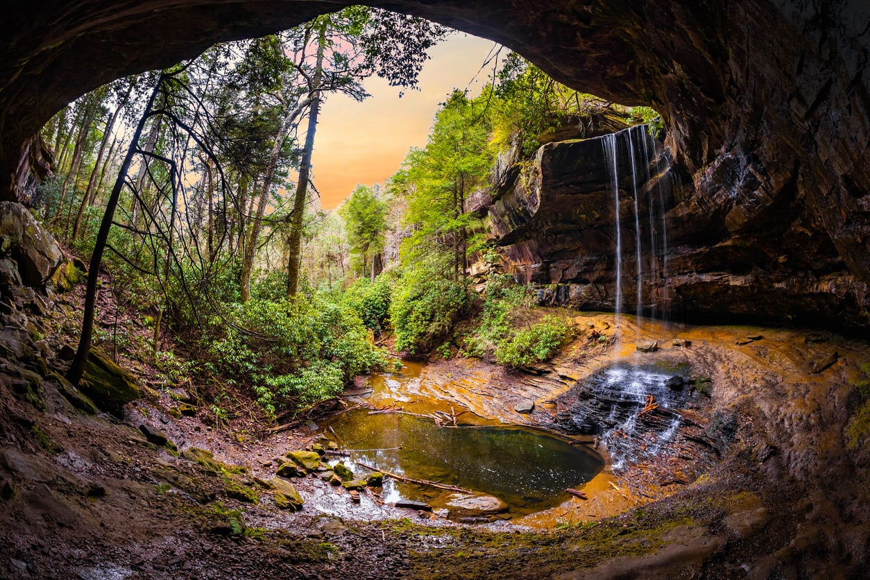 A small waterfall cascades into a shallow pool beneath a rocky overhang in a lush, green forest at sunset. Sunlight filters through the trees, illuminating mossy rocks and foliage—an idyllic scene that makes this spot one of the region’s most picturesque hiking destinations.