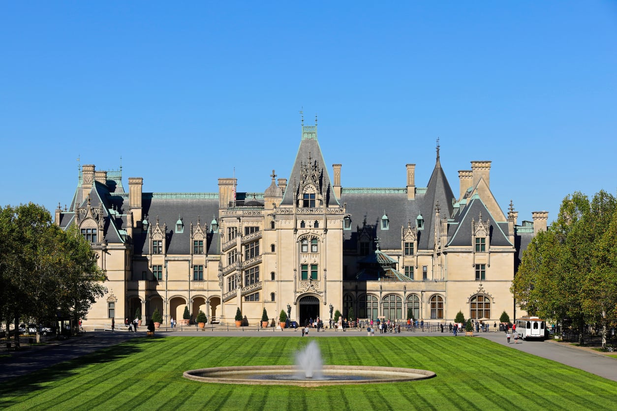 Large stone mansion with many windows and towers, reminiscent of grand castles, set behind a circular fountain and manicured lawn, surrounded by trees under a clear blue sky.