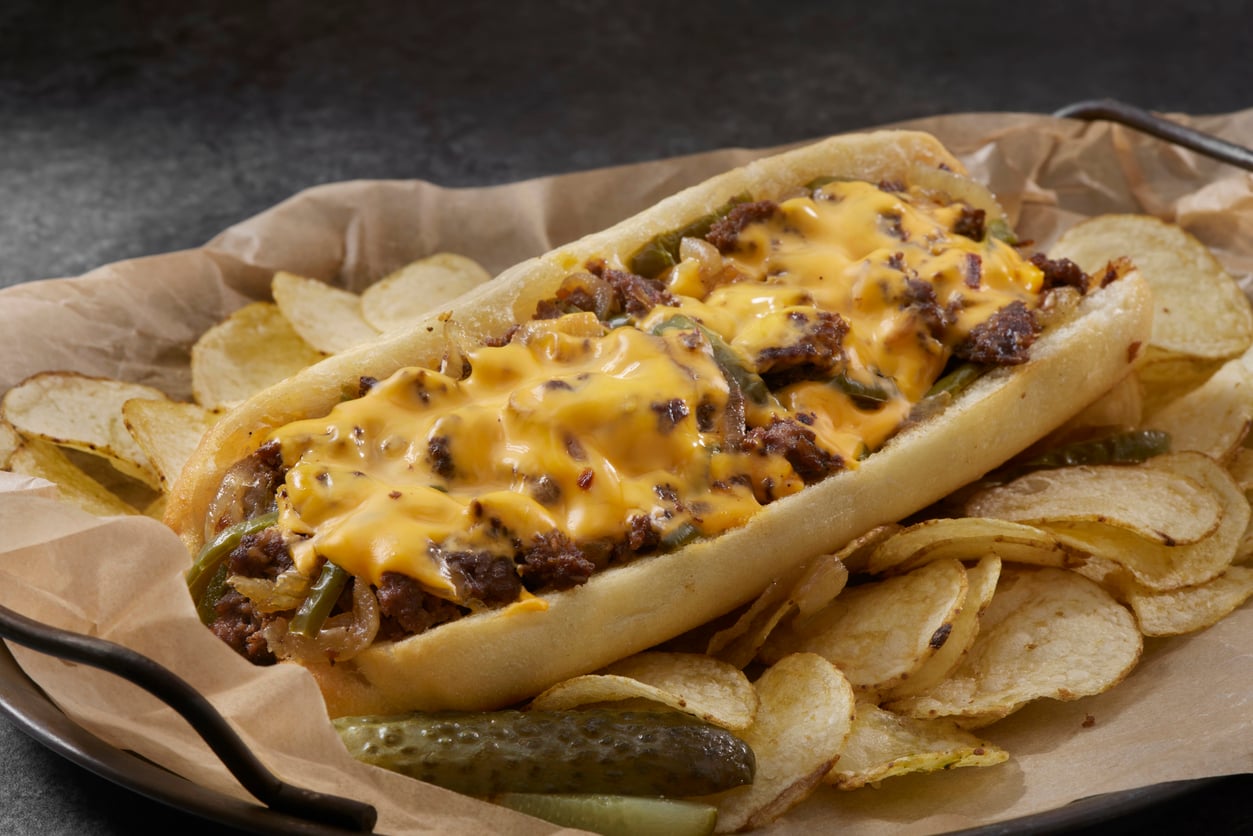 A cheesesteak sandwich topped with melted cheese sits on a tray lined with parchment paper, surrounded by potato chips and a pickle spear.