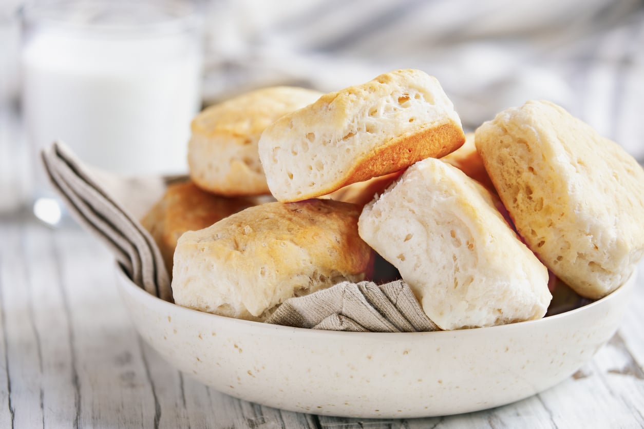 A bowl filled with golden, fluffy biscuits sits on a napkin, with a glass of milk in the blurred background on a light wooden surface.