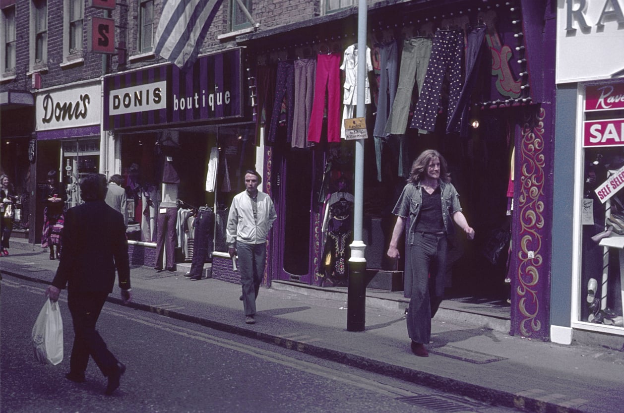 People walk along a city street lined with clothing shops. Colorful pants and shirts hang outside one store. One person carries a shopping bag. The atmosphere appears casual and urban.