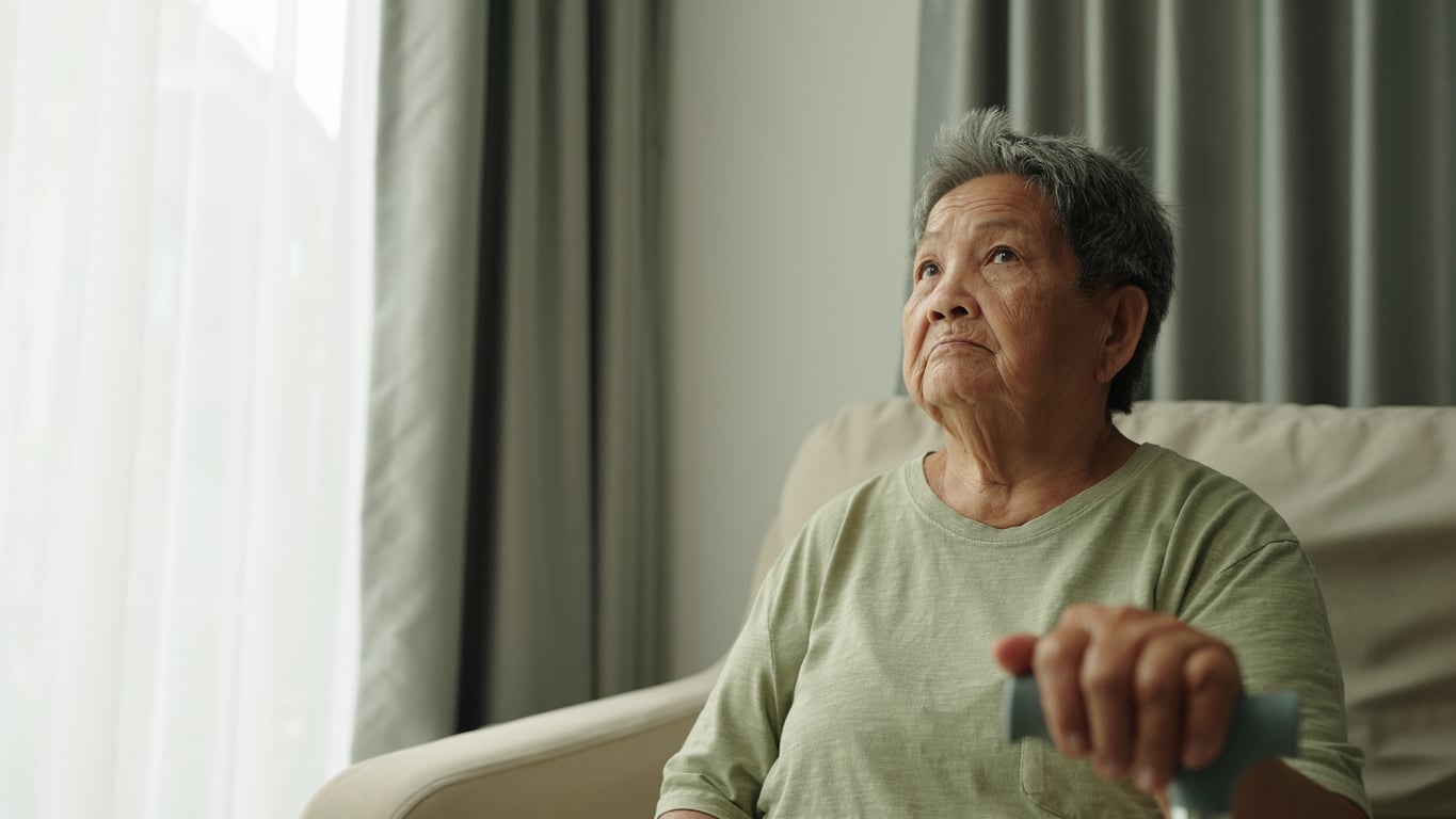 An elderly woman sits on a beige chair, looking thoughtful. She wears a light green shirt and holds a remote control, with gray curtains and sunlight through a window in the background.