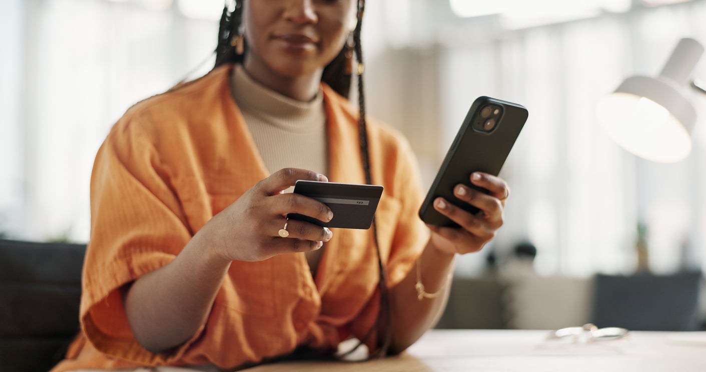 A woman wearing an orange shirt sits at a desk, holding a credit card in one hand and a smartphone in the other, appearing to make an online purchase.