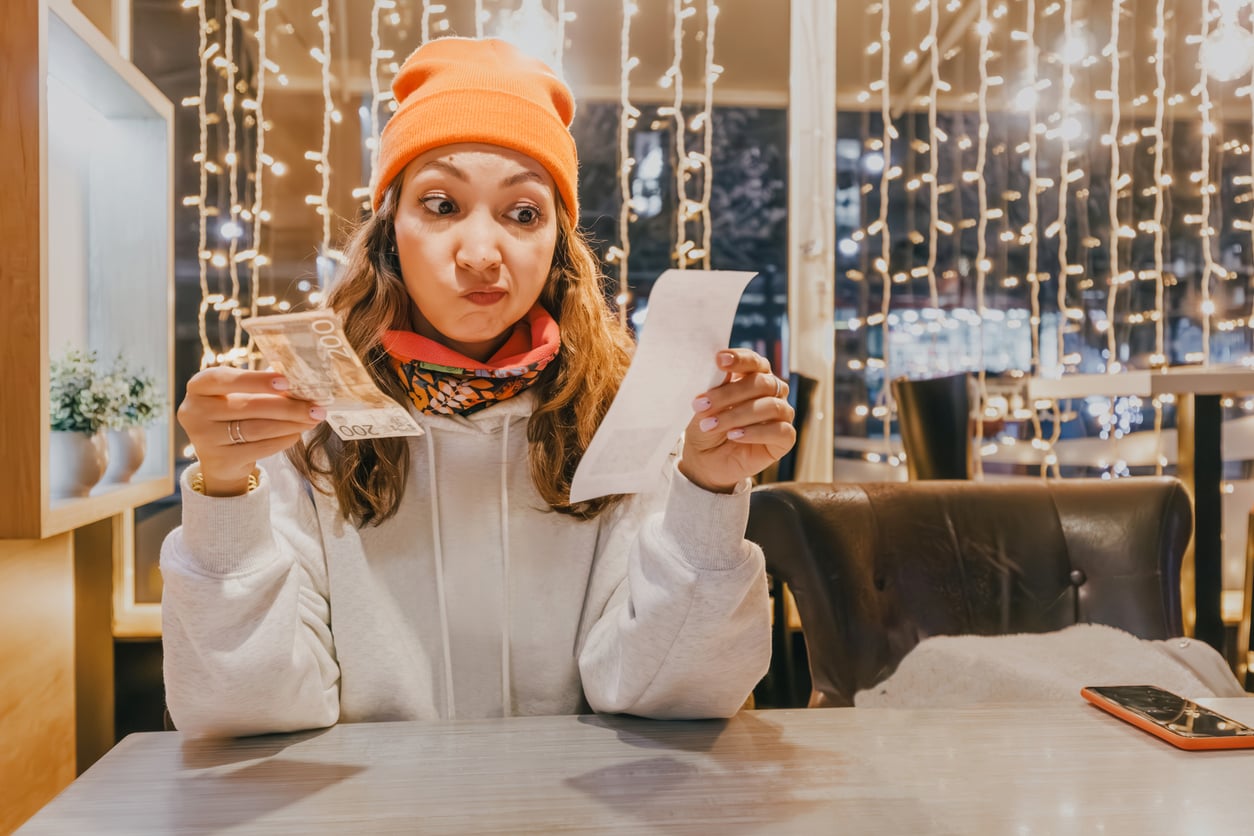 A woman in a bright orange beanie looks surprised while holding cash and a receipt at a table in a cozy, warmly lit cafe decorated with string lights.