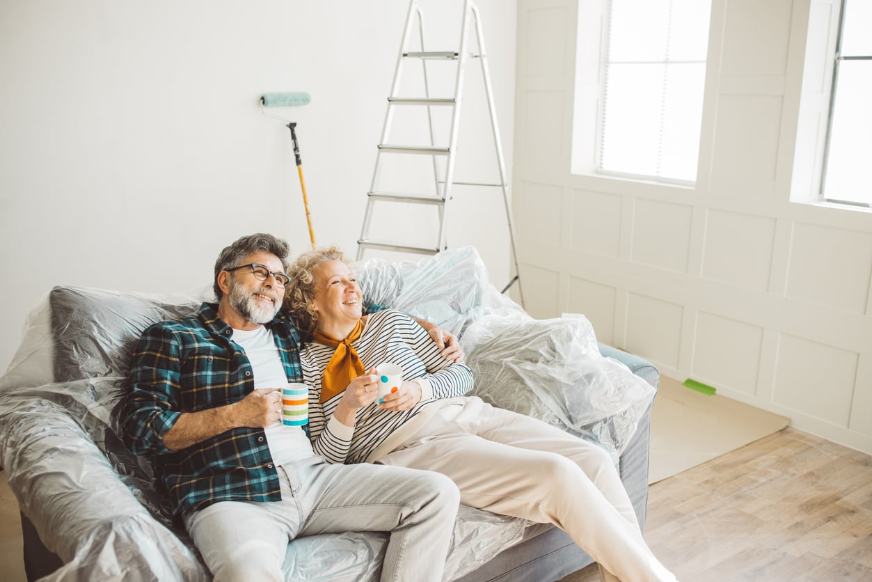 An older couple sits on a plastic-covered couch, smiling and holding mugs. Behind them are a ladder, painting supplies, and bright windows, suggesting they are renovating or redecorating their home together—perhaps finding joy in things families spend less on by tackling the project themselves.
