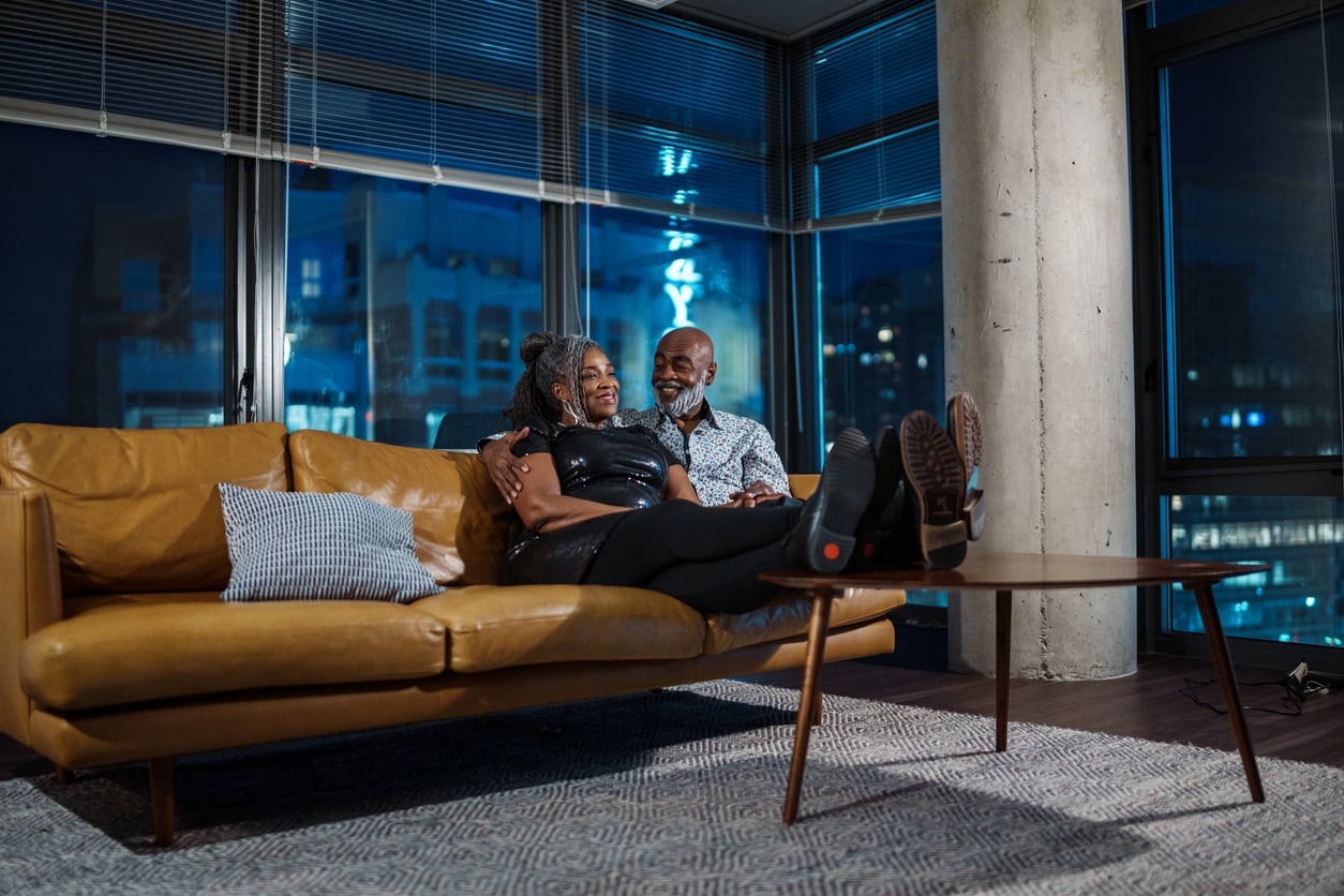 A couple relaxes on a brown sofa in a modern living room at night, smiling and leaning against each other, with city lights visible through large windows in the background.