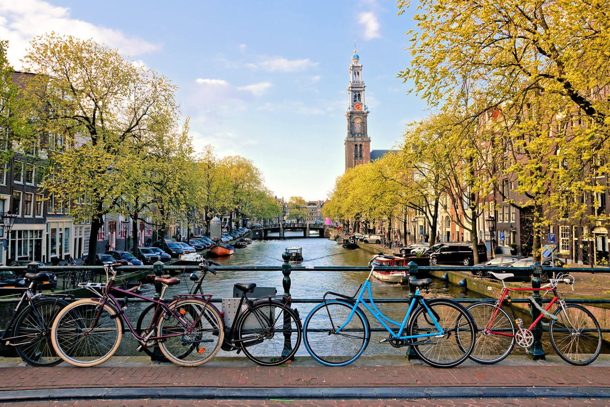 Several bicycles are parked along a bridge overlooking a canal lined with trees and buildings in Amsterdam, with a church tower visible in the background under a blue sky.