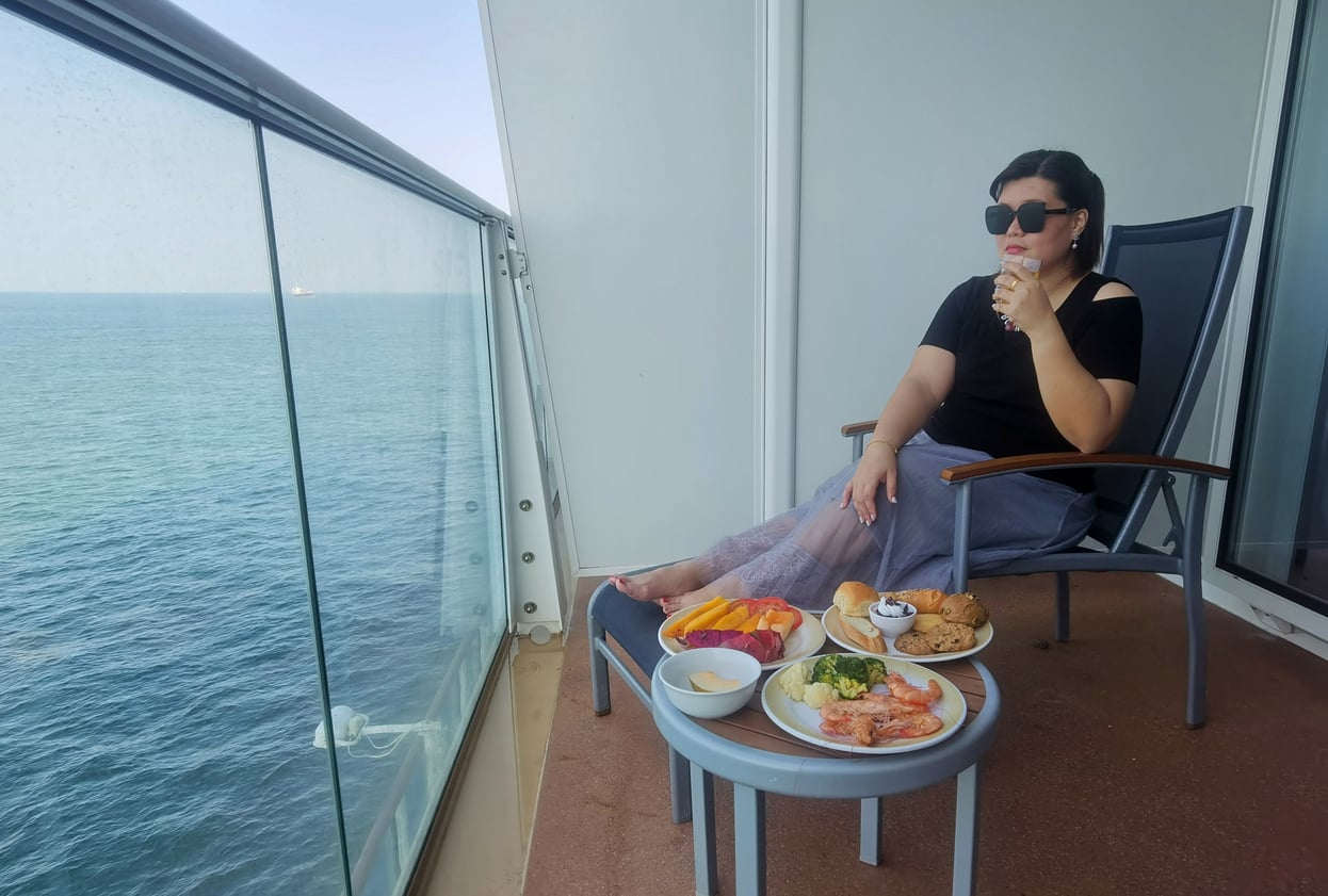 A woman sits on a balcony overlooking the ocean, wearing sunglasses and sipping a drink. A table in front of her holds fruit, croissants, and assorted food.