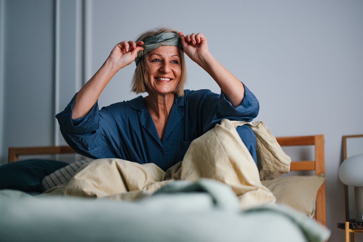 A smiling woman in blue pajamas sits on a bed, lifting a sleep mask from her eyes in a cozy, softly lit bedroom.