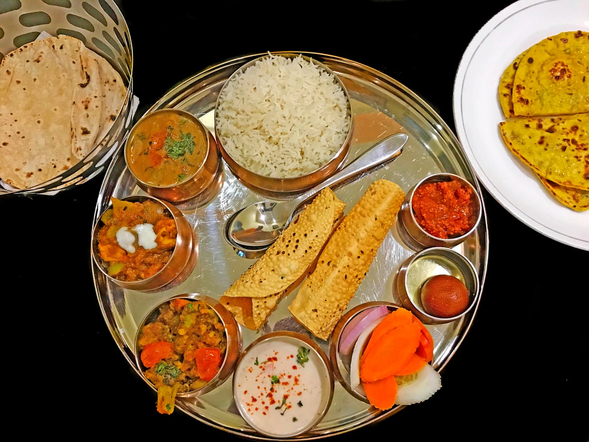 A traditional Indian thali with rice, chapati, paratha, papad, dal, vegetables, raita, pickle, salad, and a gulab jamun dessert, served on a round metal plate.