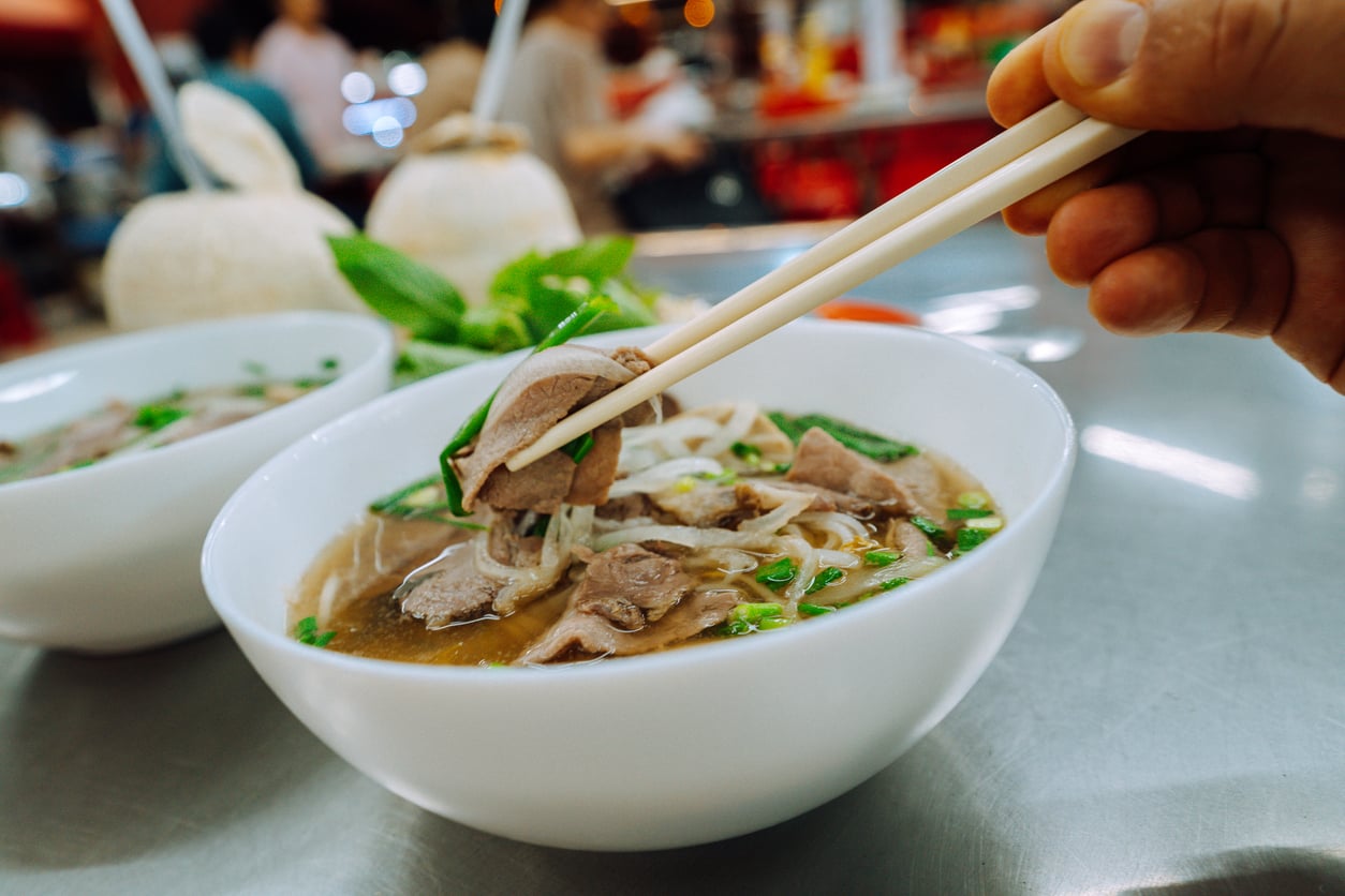 A hand uses chopsticks to lift a piece of meat from a bowl of Vietnamese pho, filled with noodles, herbs, and savory broth—one of the must-try foods to order. Another bowl and fresh herbs are in the background.