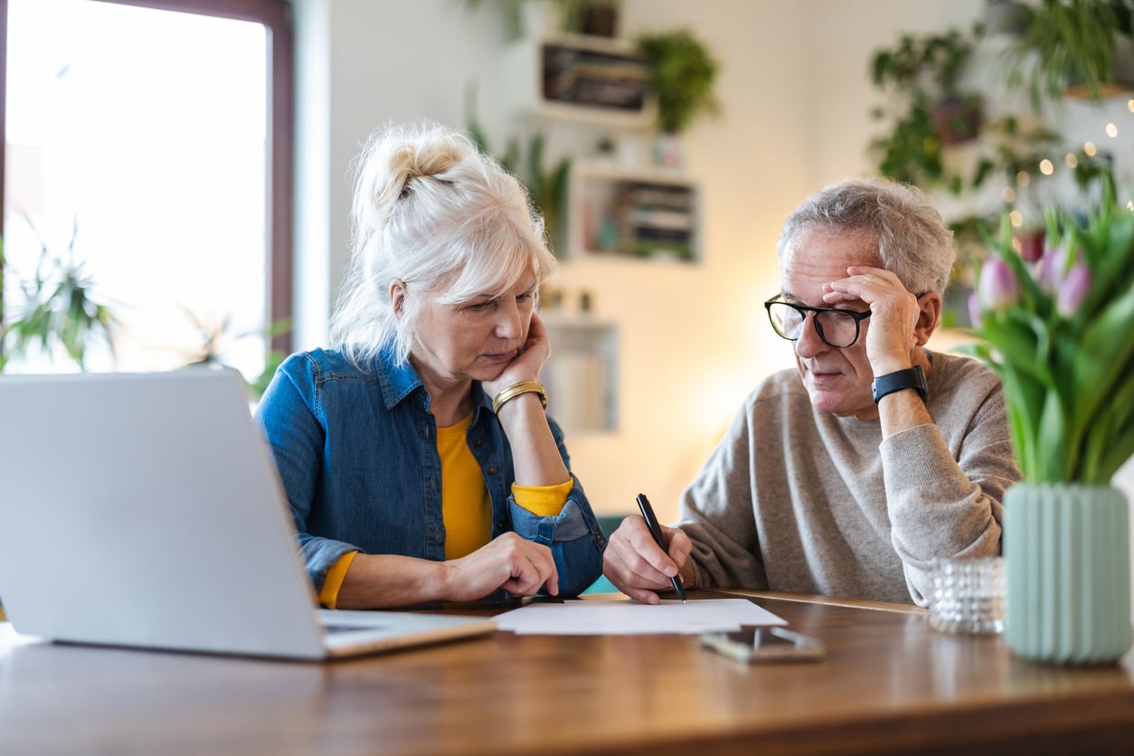 An older couple sits at a wooden table with a laptop and papers, appearing focused and concerned as they review documents related to their retirement money habits together in a bright, plant-filled room.