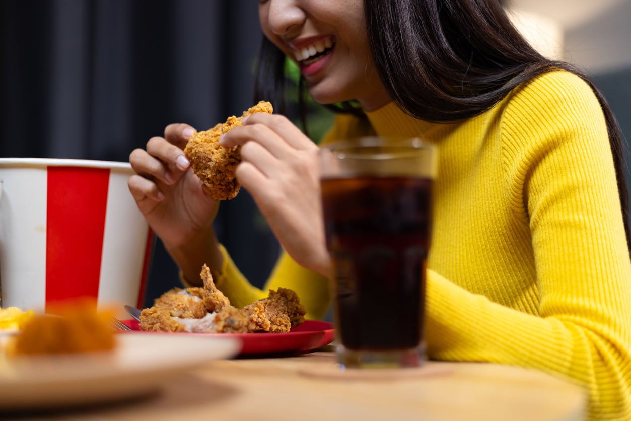 A woman in a yellow sweater smiles as she eats fried chicken, savoring one of the most popular foods to order. A glass of dark soda and a bucket of chicken are on the table in front of her.
