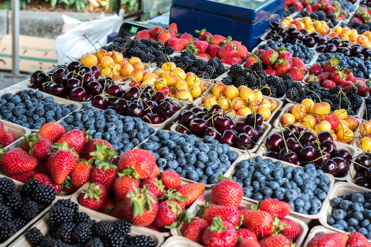 A market stall displays baskets of fresh berries, including strawberries, blueberries, blackberries, red cherries, yellow cherries, and raspberries arranged in neat rows.