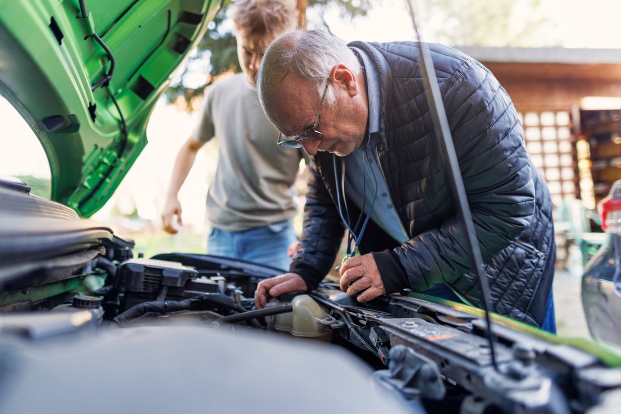 An older man in a black jacket examines a car engine under an open hood, while a younger person stands nearby, assisting him outside on a sunny day.