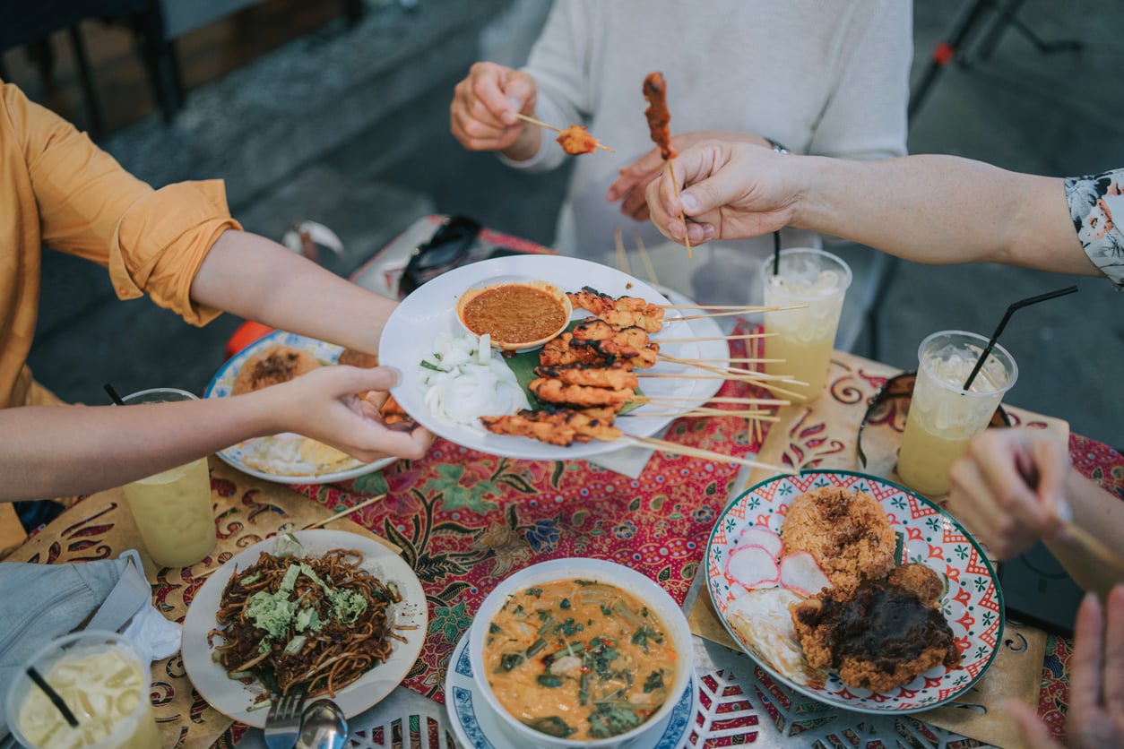 Four people seated around a table filled with various dishes, including skewered grilled meat, curry, noodles, and iced drinks, with hands reaching for food.