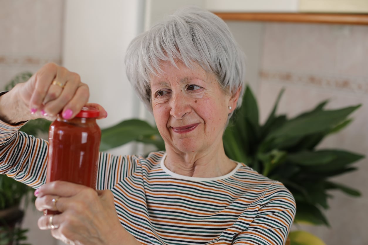 An older woman with short gray hair and a striped shirt smiles while opening a jar of red sauce in a kitchen with green plants in the background.