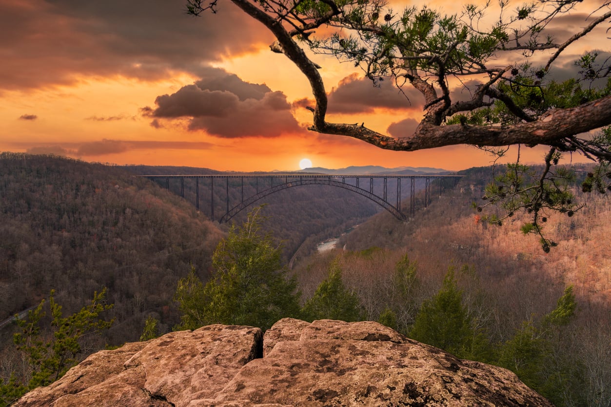A scenic sunset view of a steel arch bridge spanning a forested valley—one of the region’s most inspiring hiking destinations—with a rocky outcrop and pine tree branches in the foreground and the sun setting behind the hills.