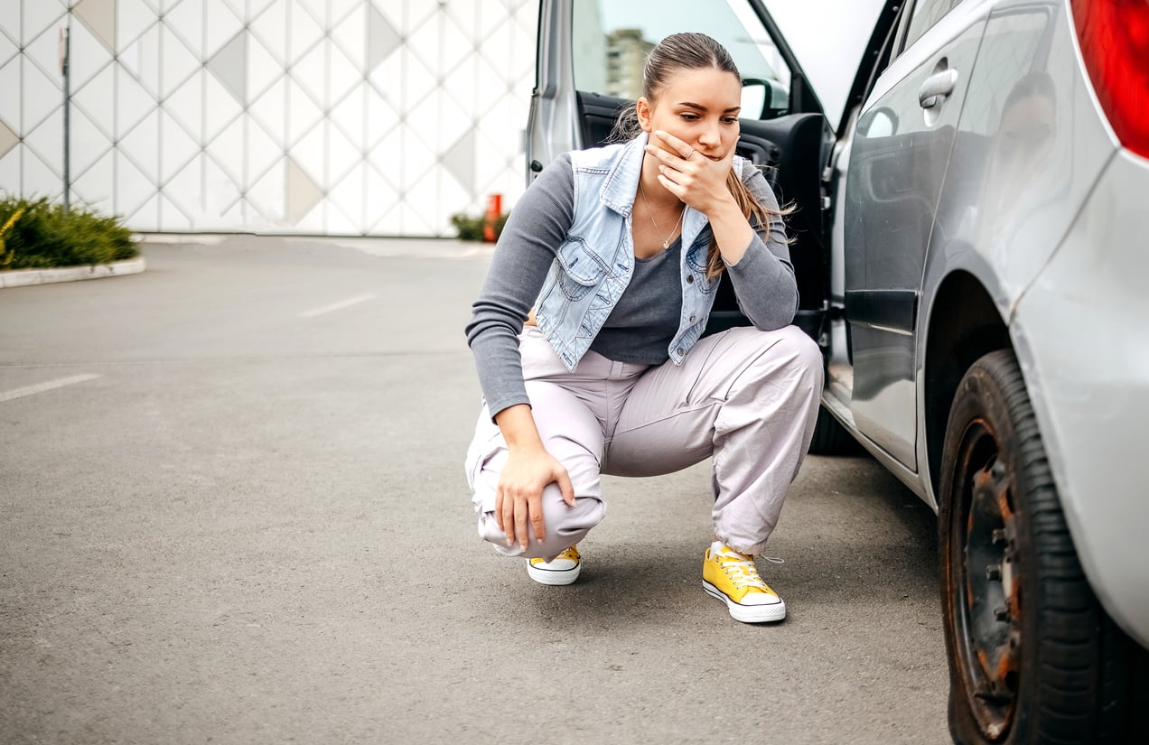 A person crouches next to a car with a flat tire, covering their mouth with one hand and looking concerned.