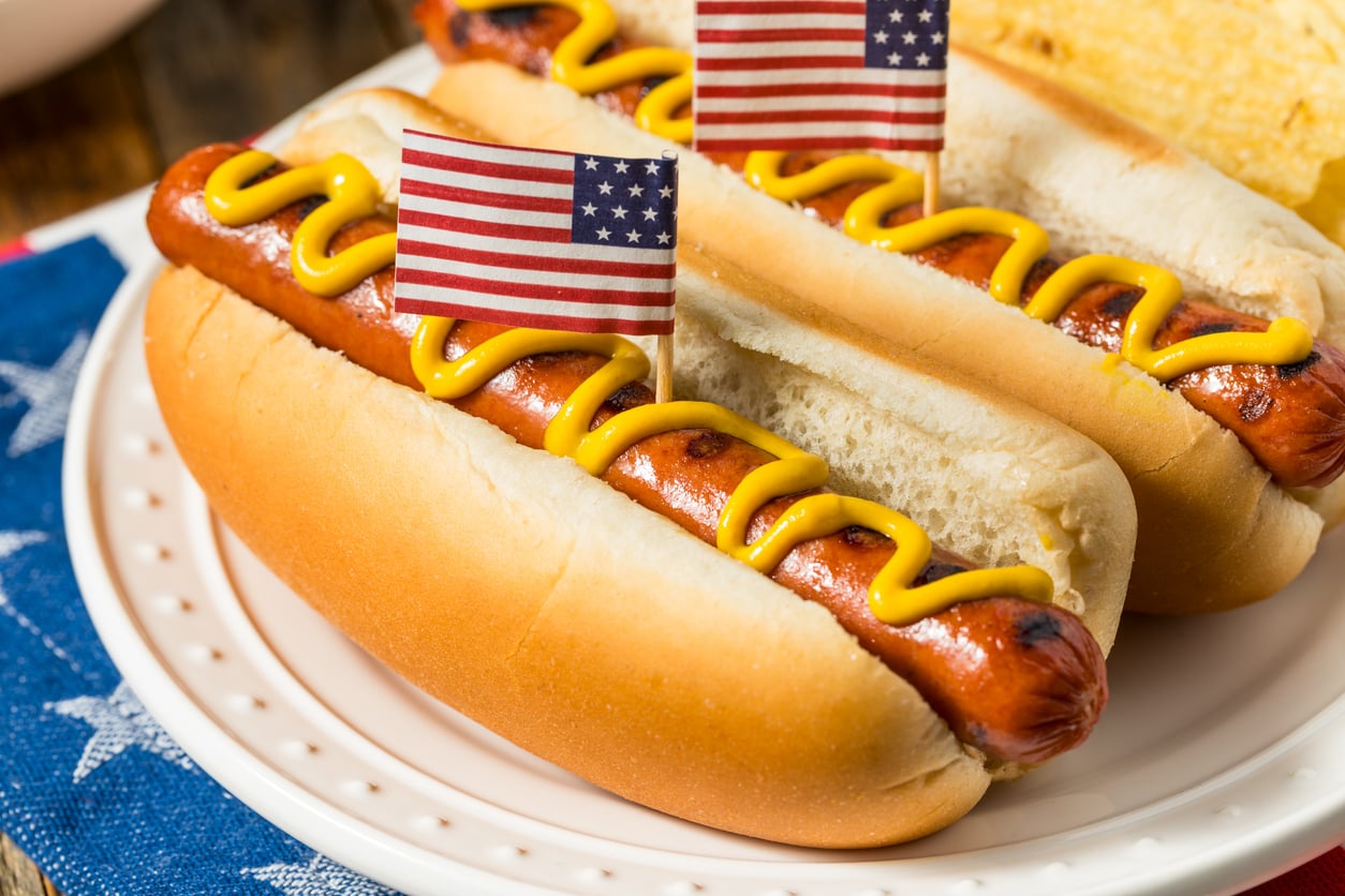 Two hot dogs in buns topped with mustard and small American flags on a plate, with part of a blue star-patterned cloth visible underneath.