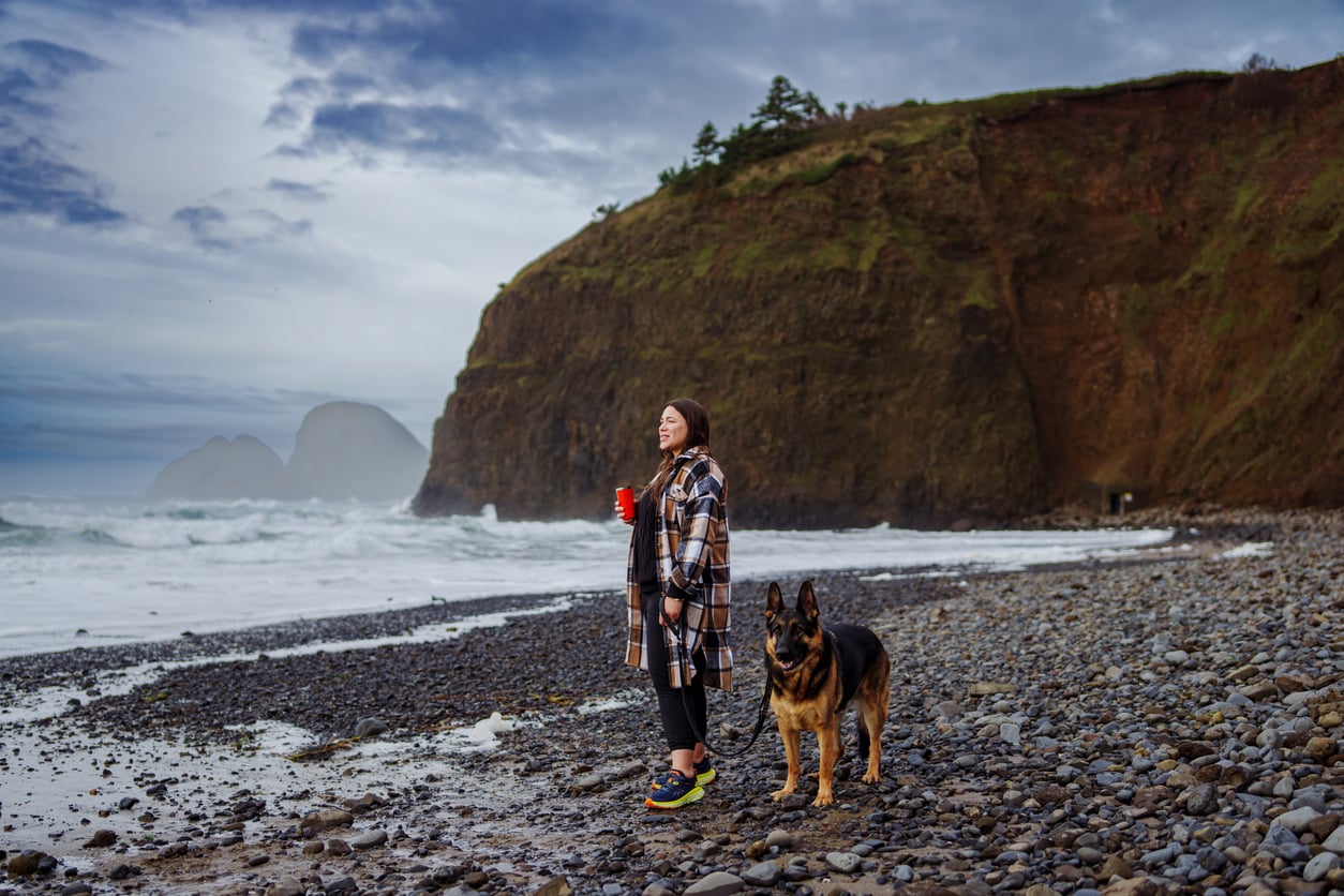 A woman holding a red cup stands on a rocky beach beside a German Shepherd, with cliffs and ocean waves in the background under a cloudy sky.
