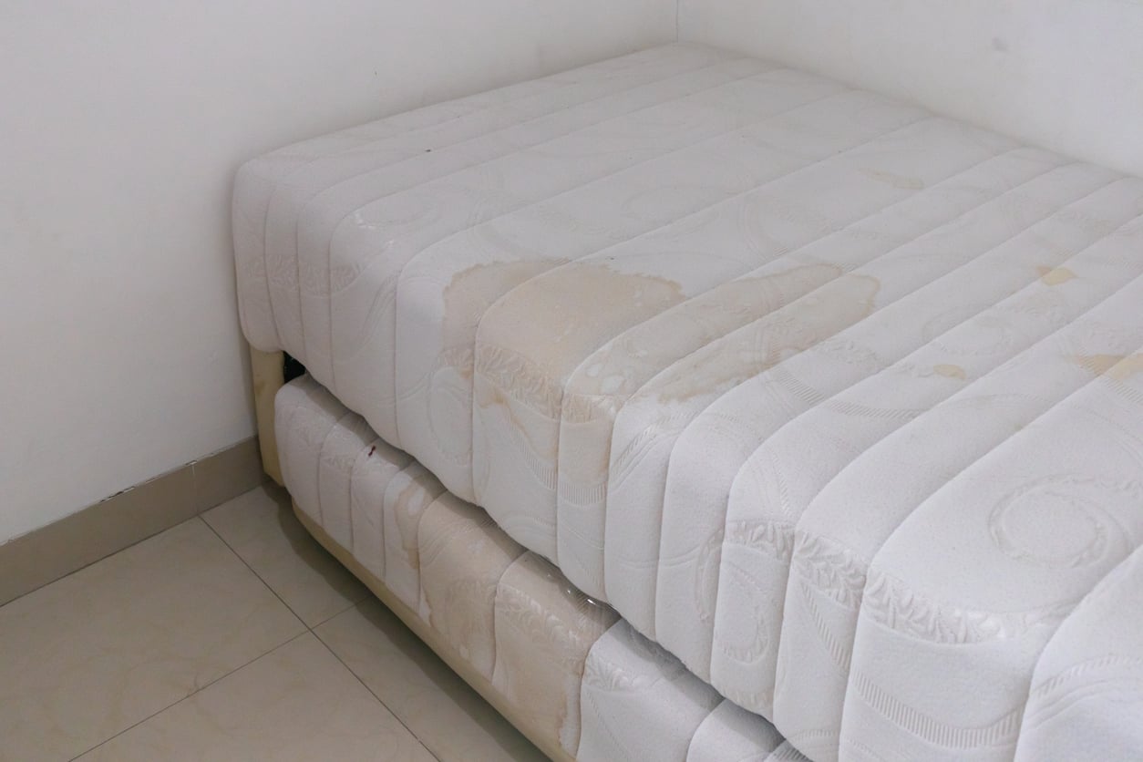 Two white mattresses with visible stains are stacked in the corner of a room with tiled flooring and white walls.