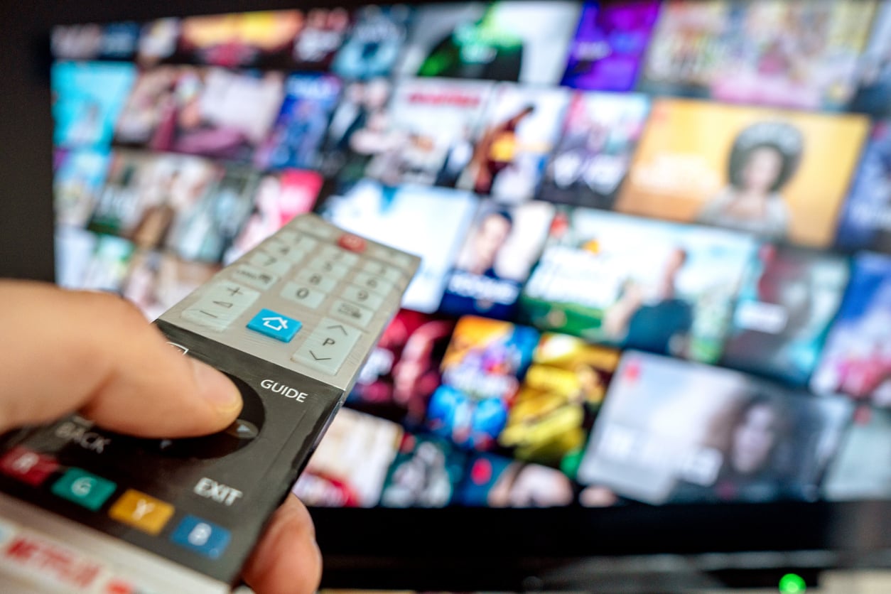 A hand holds a TV remote pointed at a television screen displaying a colorful selection of streaming service thumbnails and shows in a grid layout, slightly out of focus.