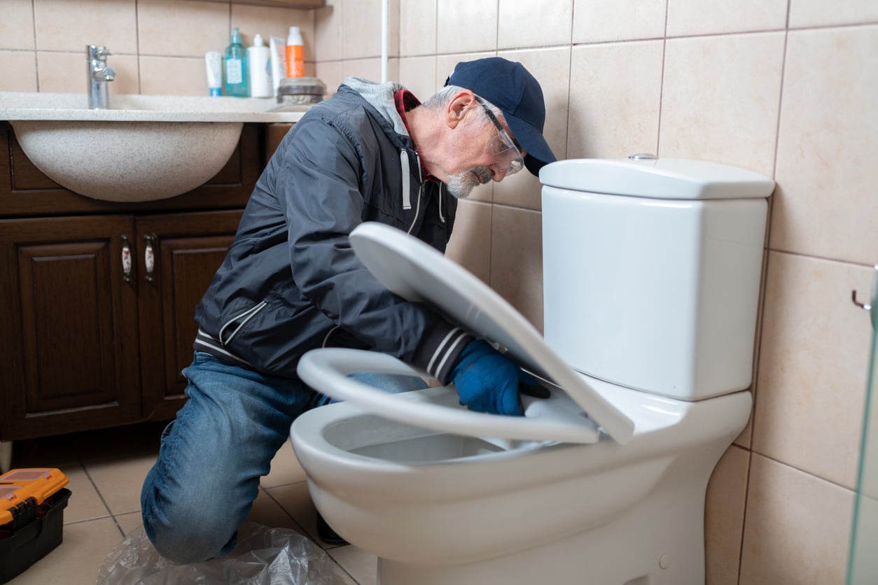 An older man wearing a cap and gloves kneels in a bathroom while repairing or inspecting a toilet, with tools and cleaning supplies visible nearby.