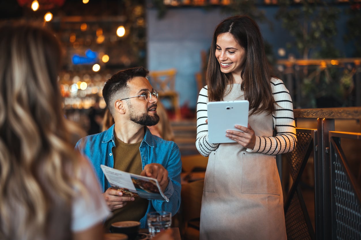 A smiling waitress holding a tablet takes an order from a seated man holding a menu in a cozy, warmly lit restaurant.