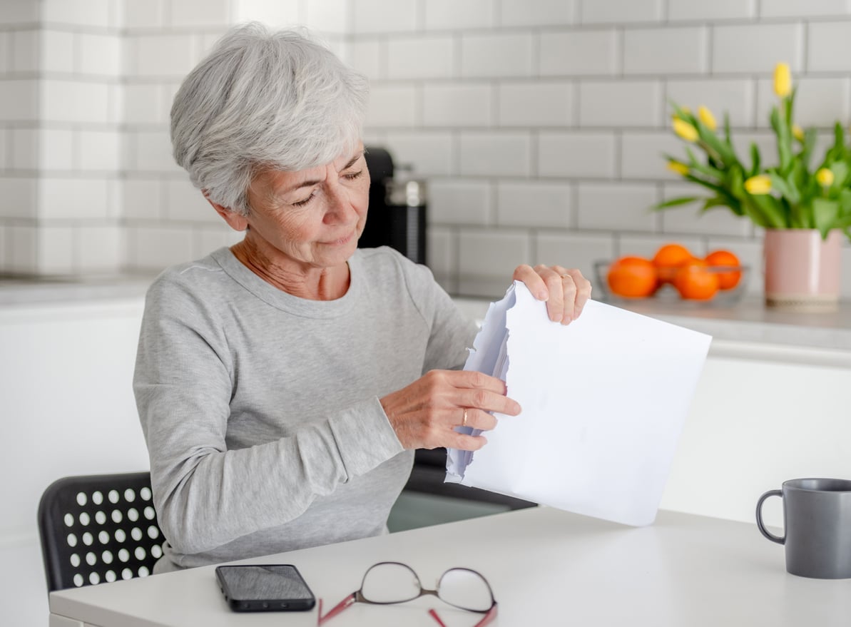 An older woman with gray hair sits at a kitchen table, tearing a sheet of paper. A smartphone, glasses, a coffee mug, and a vase with yellow tulips are on the table.