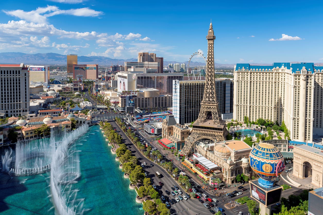 Aerial view of the Las Vegas Strip featuring the Eiffel Tower replica at Paris Las Vegas, hotels, casinos, fountains, and busy streets under a bright blue sky.