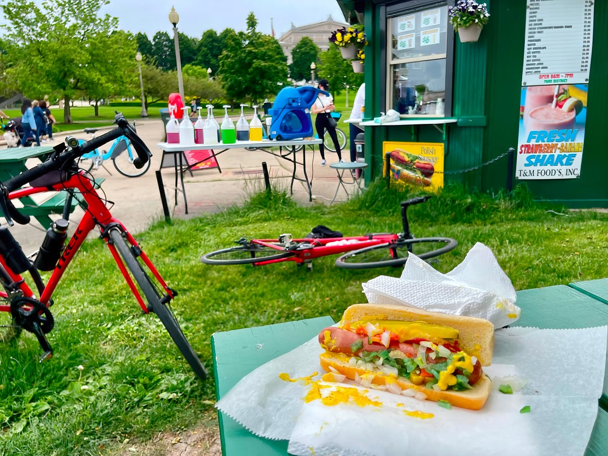 A Chicago-style hot dog on a table, with a bike lying on grass nearby, a food stand in the background, and colorful drink syrups on a table outside.