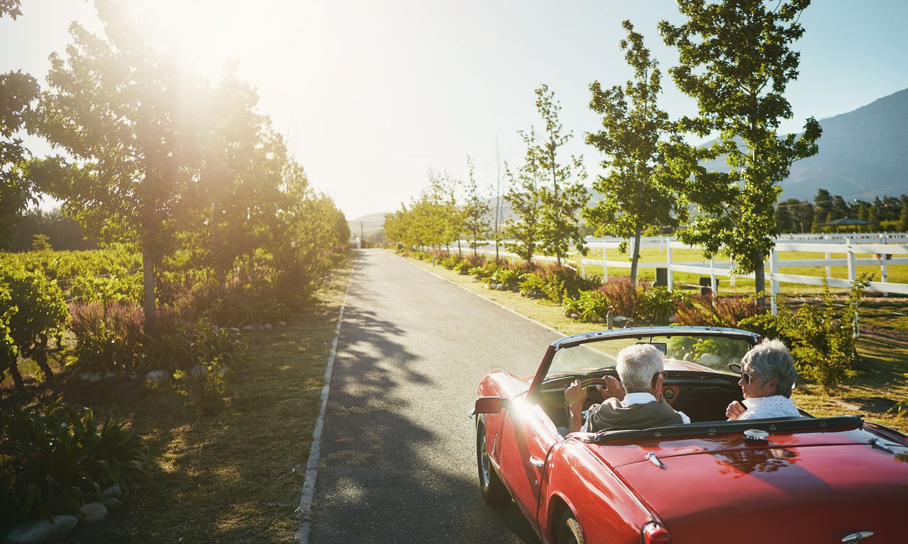 An older couple enjoys unforgettable vacation experiences as they drive a red convertible down a sunny, tree-lined rural road with open fields and mountains in the background.